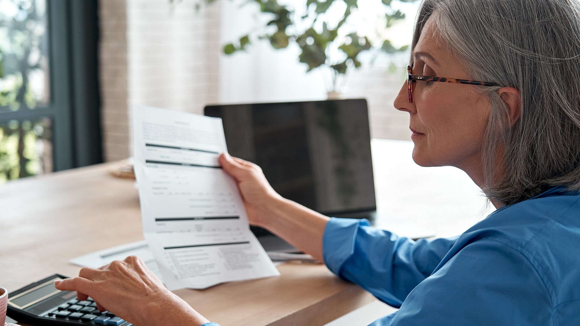 Mature woman holding paper bill using calculator managing finances at home.